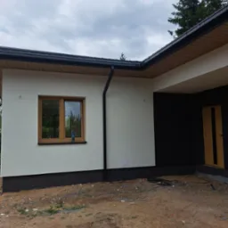 Nowoczesny dom jednorodzinny z jasną elewacją i ciemnymi akcentami, widoczne okno z brązową ramą i fragment dachu. Teren wokół domu w stanie przygotowania.