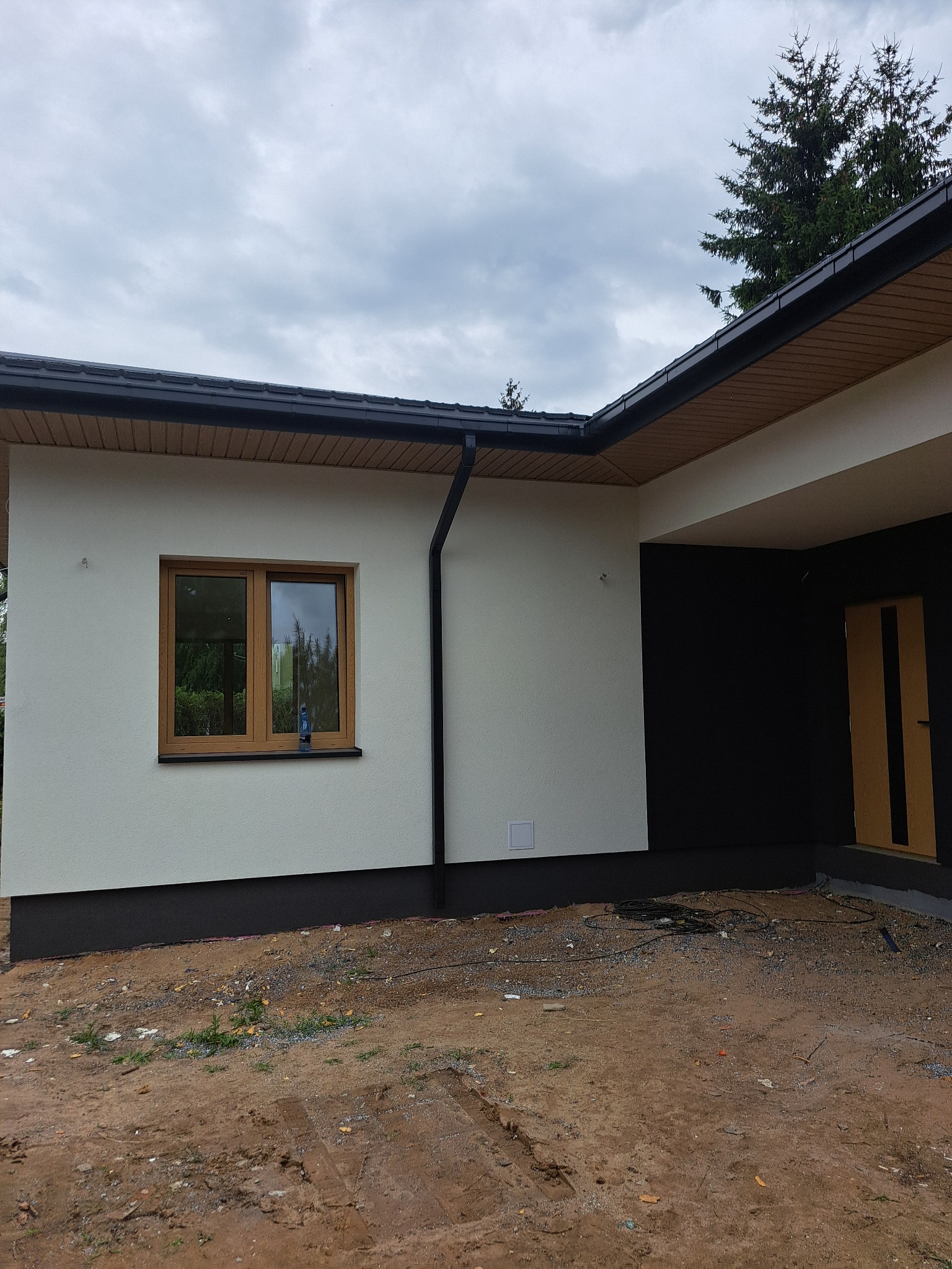 Nowoczesny dom jednorodzinny z jasną elewacją i ciemnymi akcentami, widoczne okno z brązową ramą i fragment dachu. Teren wokół domu w stanie przygotowania.