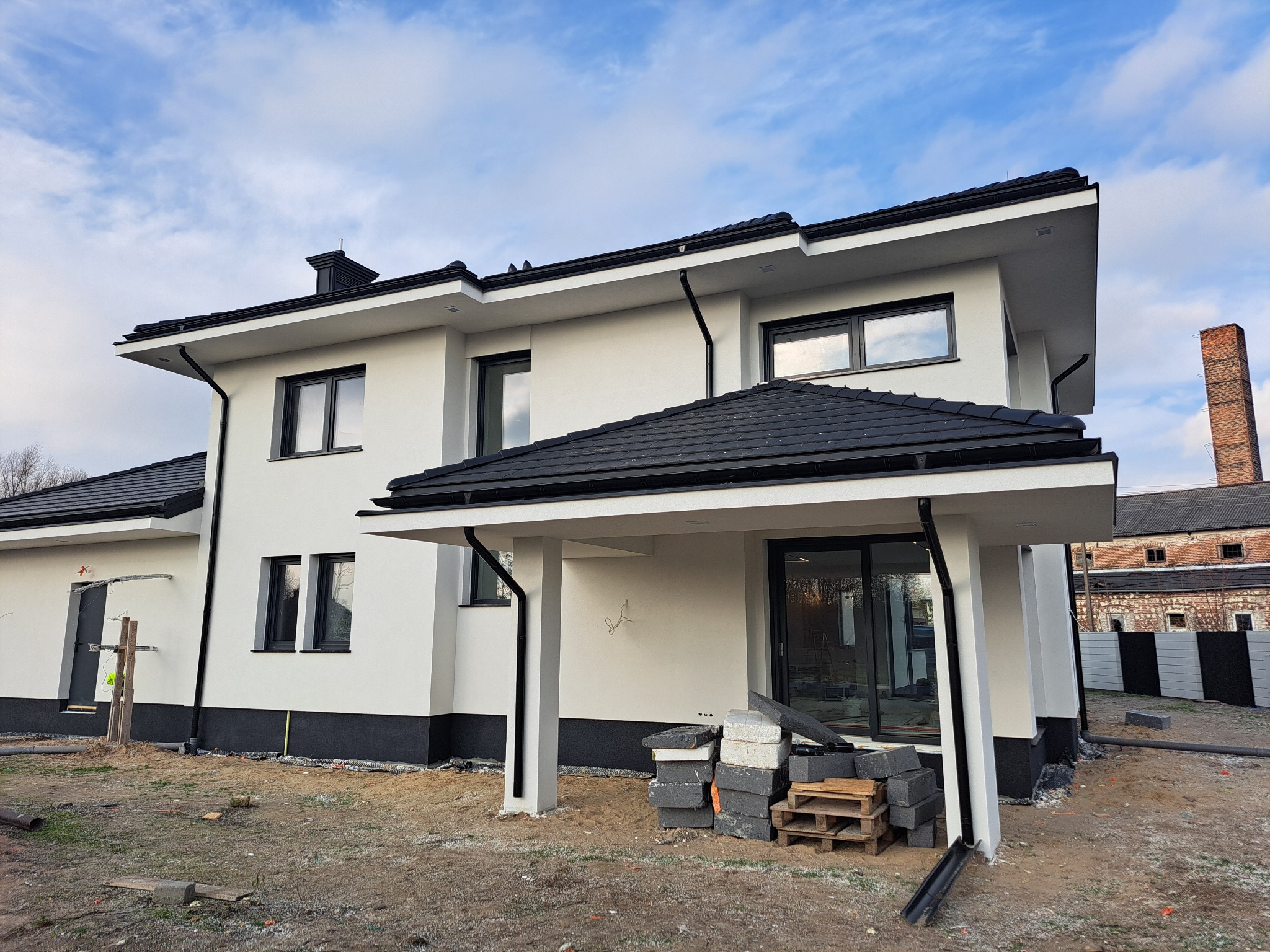 Nowoczesny, dwukondygnacyjny dom z białą elewacją i czarnym dachem. Widoczne materiały budowlane sugerują prace wykończeniowe. W tle ceglany budynek.