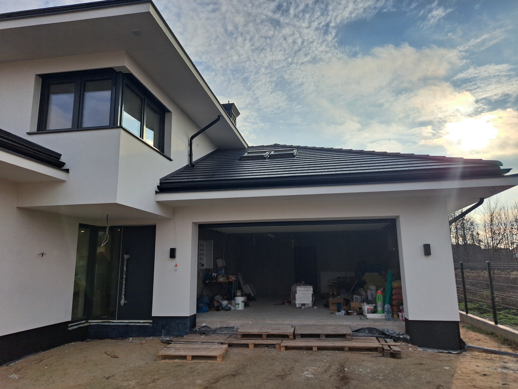 Nowoczesny dom z białą elewacją i czarnymi detalami, widok na garaż z paletami na zewnątrz. Kontrastowe kolory, błękitne niebo z chmurami w tle.