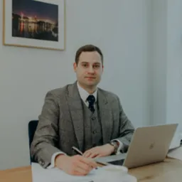 Mężczyzna w tweedowej marynarce i kamizelce siedzi przy biurku, pisząc na papierze. Na biurku laptop, filiżanka kawy i zdjęcie w ramce na ścianie.