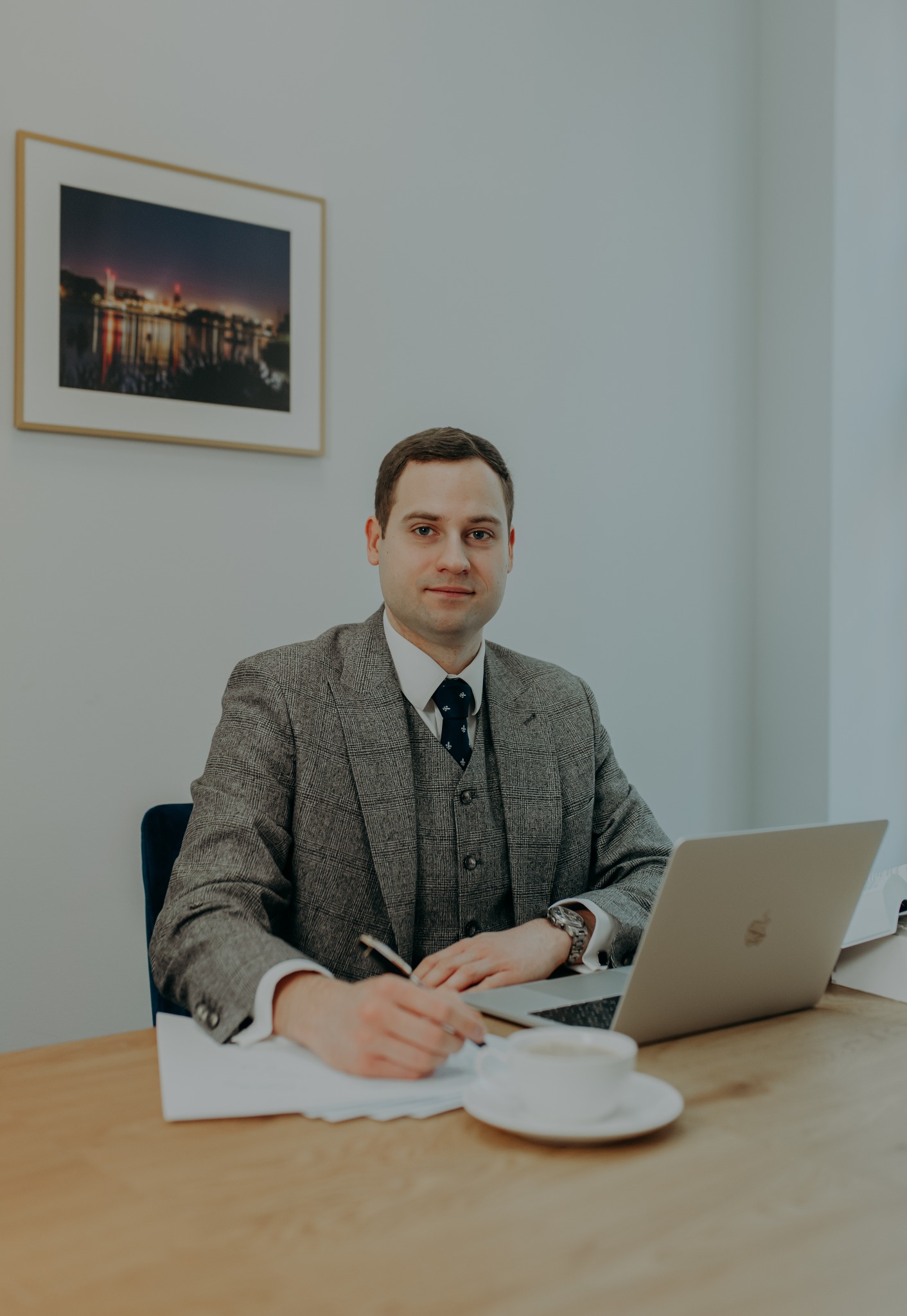 Mężczyzna w tweedowej marynarce i kamizelce siedzi przy biurku, pisząc na papierze. Na biurku laptop, filiżanka kawy i zdjęcie w ramce na ścianie.