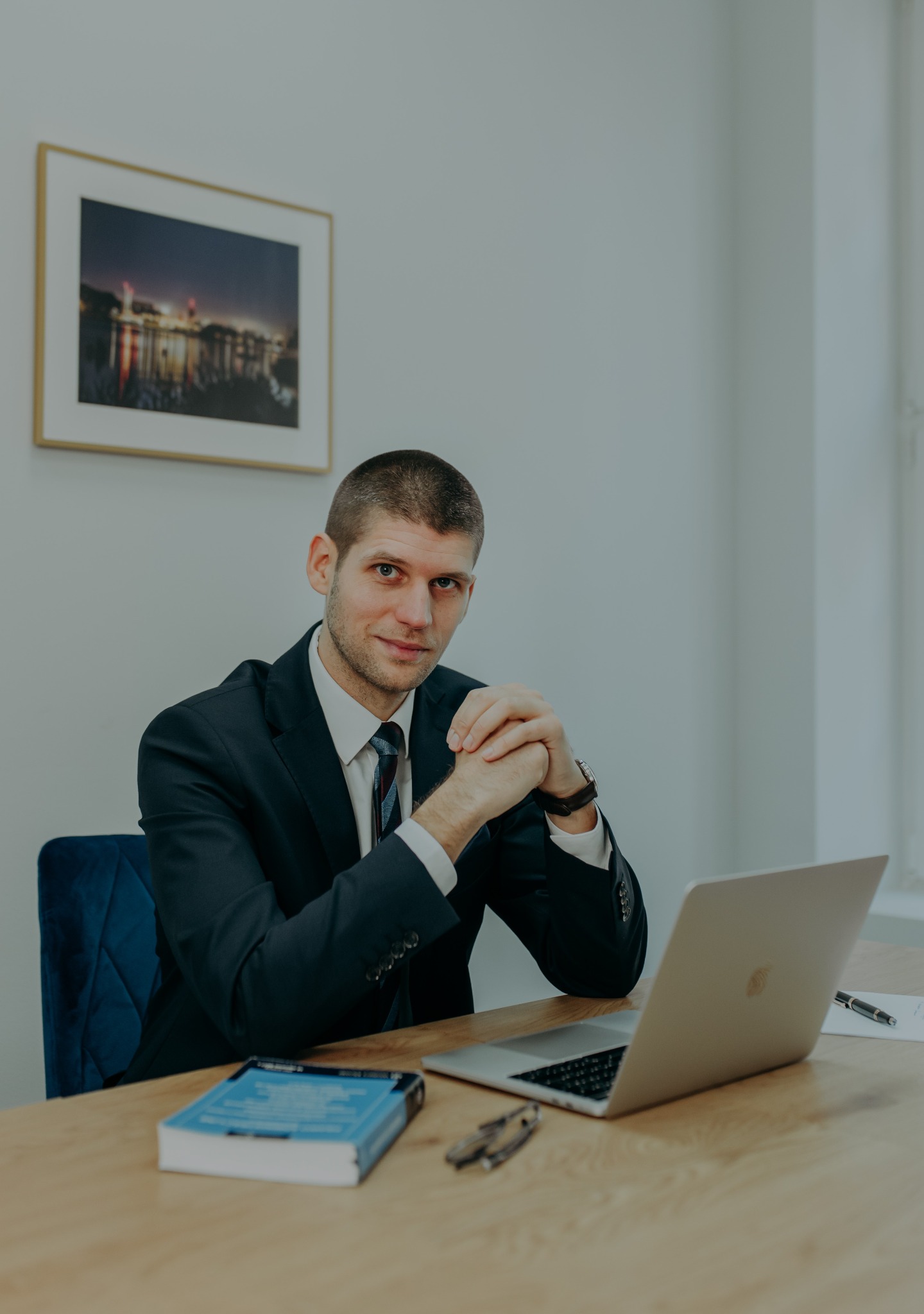 Portret prawnika w garniturze siedzącego za biurkiem z laptopem, książką prawniczą i okularami, w tle obraz z motywem miejskim.