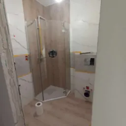 Montaż kabiny prysznicowej w łazience z imitacją drewna na ścianach i podłodze, widoczne niedokończone prace wykończeniowe przy ścianie i instalacji WC.