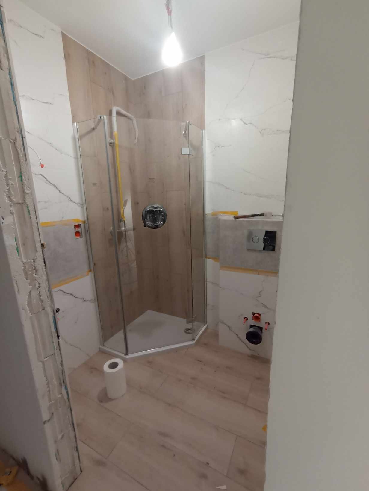 Montaż kabiny prysznicowej w łazience z imitacją drewna na ścianach i podłodze, widoczne niedokończone prace wykończeniowe przy ścianie i instalacji WC.