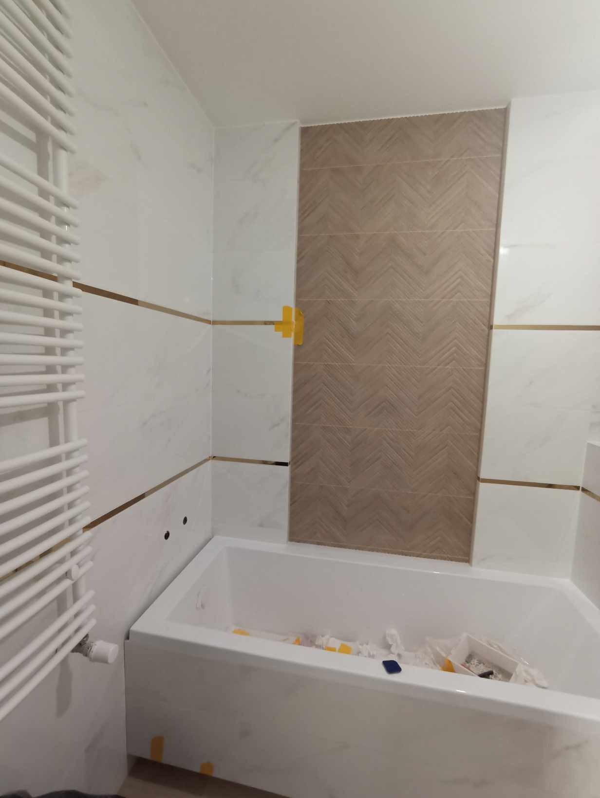 Biała wanna w łazience z białymi płytkami imitującymi marmur, oddzielonymi złotymi paskami, oraz dekoracyjnym panelem z płytek w odcieniach brązu, ułożonych w jodełkę; widoczne żółte krzyżyki...