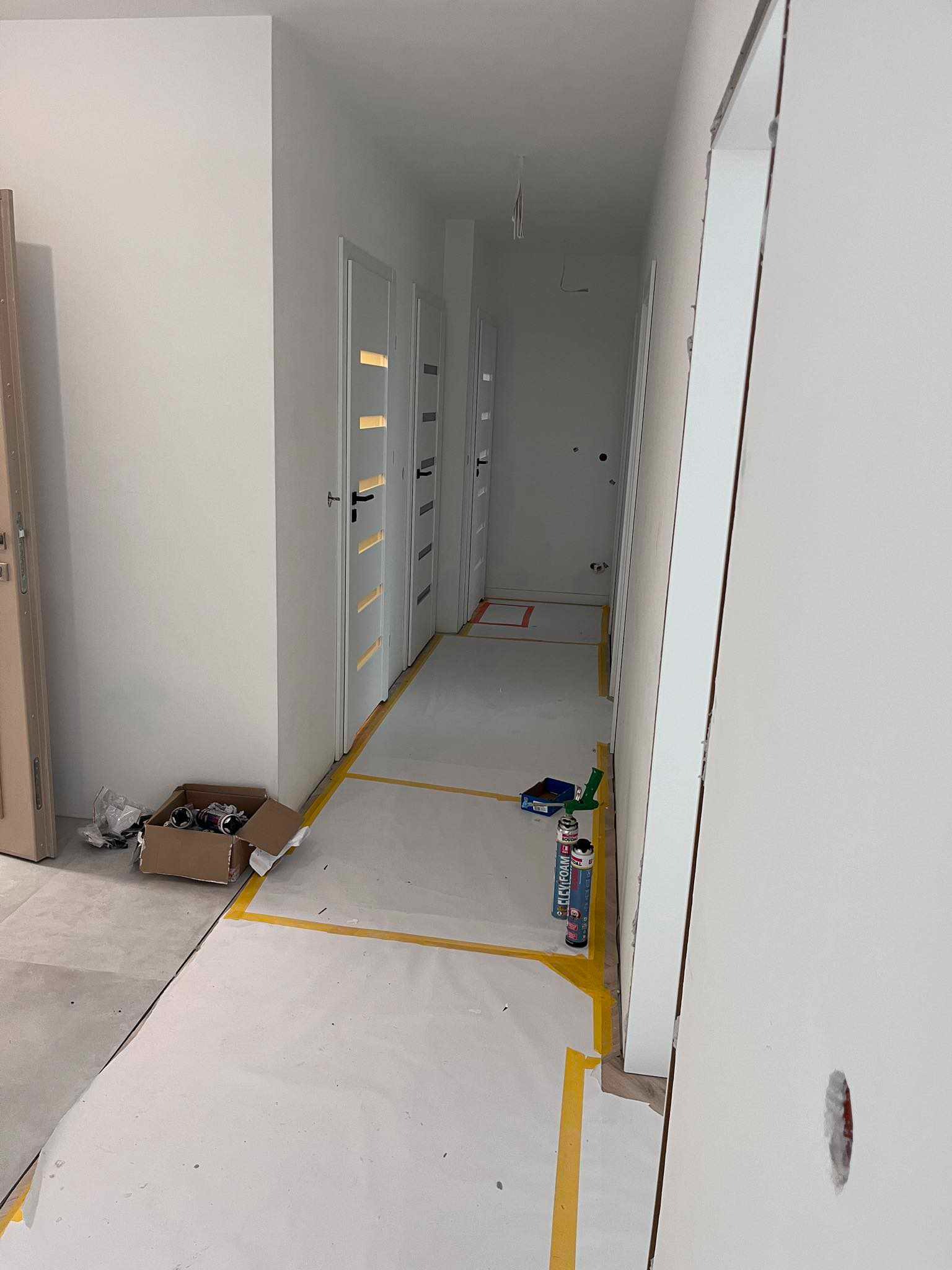 Korytarz w trakcie prac wykończeniowych: zabezpieczona podłoga folią i taśmą, puszki z pianką montażową, widoczne trzy białe drzwi z poziomymi przeszkleniami i czarnymi klamkami, karton...