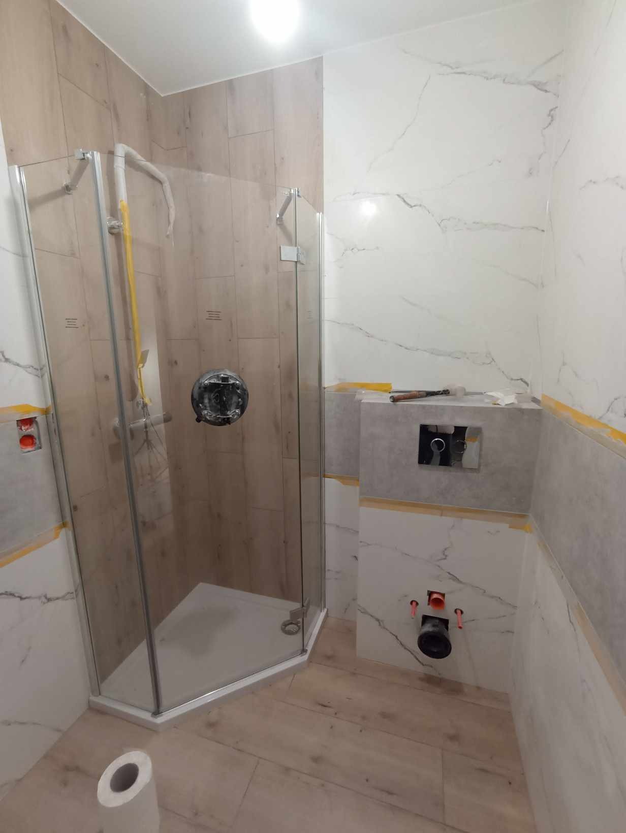 Montaż kabiny prysznicowej z elementami instalacji wodnej i częściowo wykończonymi ścianami z płytek imitujących marmur i drewno, widoczne zabezpieczenia taśmą malarską.