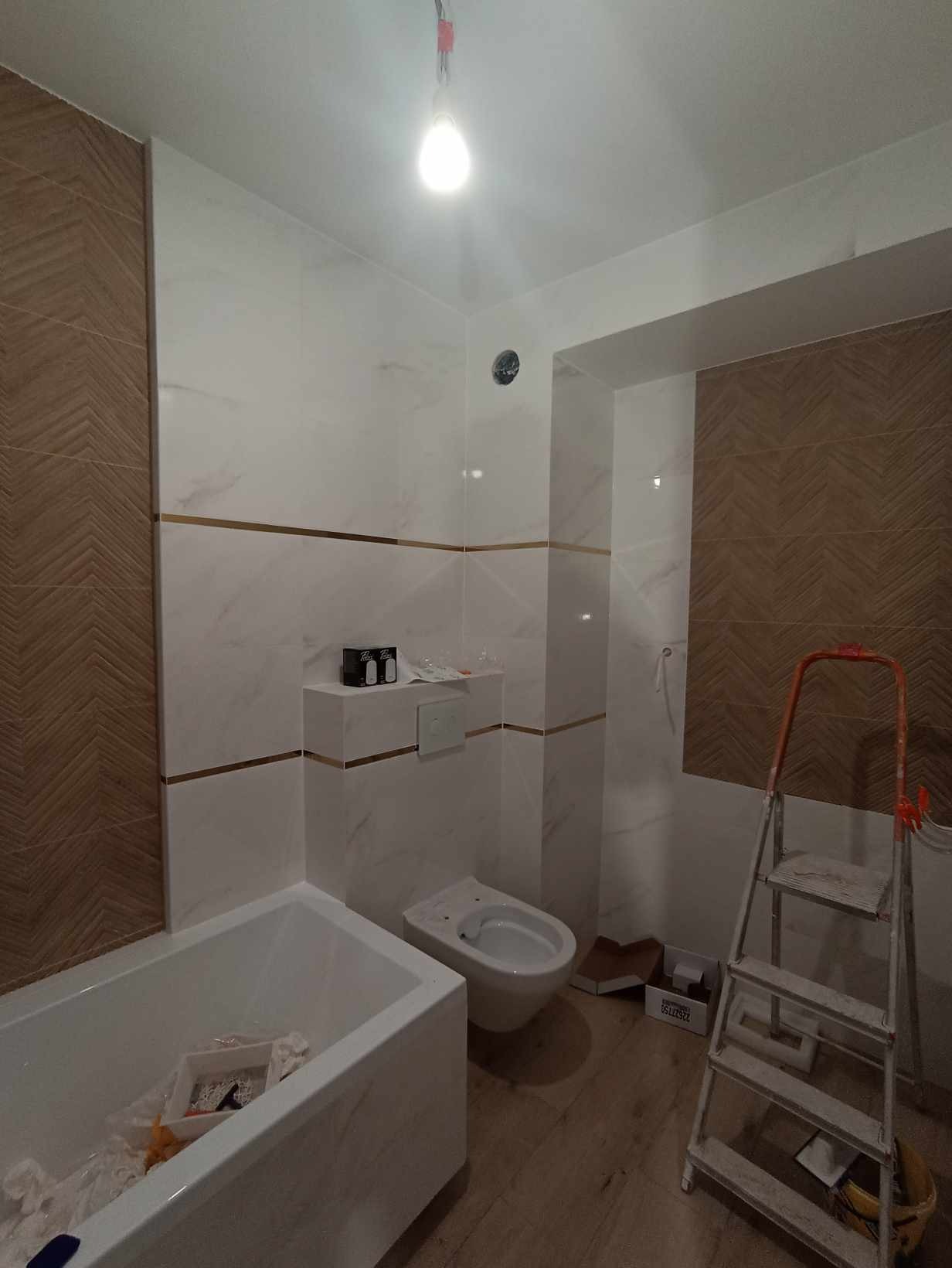 Wykończona łazienka z białymi i brązowymi płytkami, złotymi paskami dekoracyjnymi, białą wanną i toaletą, widoczna drabina malarska, podłoga drewnopodobna.