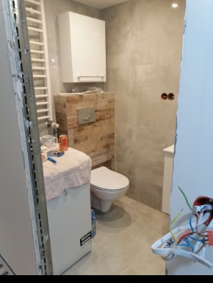 Wnętrze łazienki w trakcie remontu: widoczne płytki imitujące beton, zabudowa WC z drewnopodobnymi panelami, biała szafka wisząca i grzejnik drabinkowy, instalacja elektryczna wystająca ze ściany.