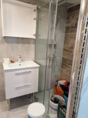 Nowoczesna łazienka z białą szafką pod umywalką, kabiną prysznicową ze szklanymi drzwiami i widocznymi narzędziami budowlanymi w tle, ukazującymi etap prac wykończeniowych.