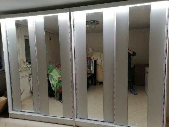 Szafa przesuwna z oświetleniem LED w górnej części, widoczne odbicie pomieszczenia i osoby w lustrzanych drzwiach.