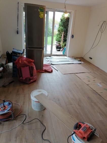 Przygotowanie do montażu paneli podłogowych: widoczna piła do paneli, wiadro z klejem, rozłożone panele i przewody elektryczne wystające ze ściany.