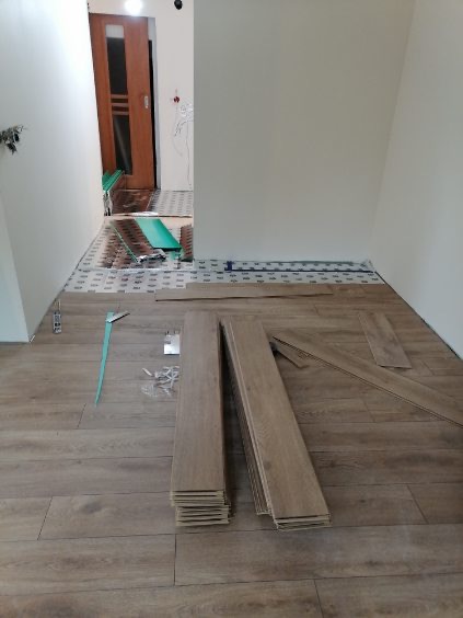 Układanie paneli podłogowych w jasnym pomieszczeniu, widoczne rozłożone panele, podkład i narzędzia montażowe. Na ścianie widoczne wystające przewody elektryczne.