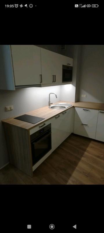 Nowoczesna kuchnia w kształcie litery L z białymi szafkami i drewnianym blatem, oświetleniem podszafkowym LED i płytą indukcyjną, widok na zlew i baterię.