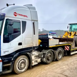 Ciężarówka laweta marki Iveco z żółtą ładowarką kołową na platformie, widoczne logo firmy transportowej Transal, w tle inne pojazdy na placu budowy.