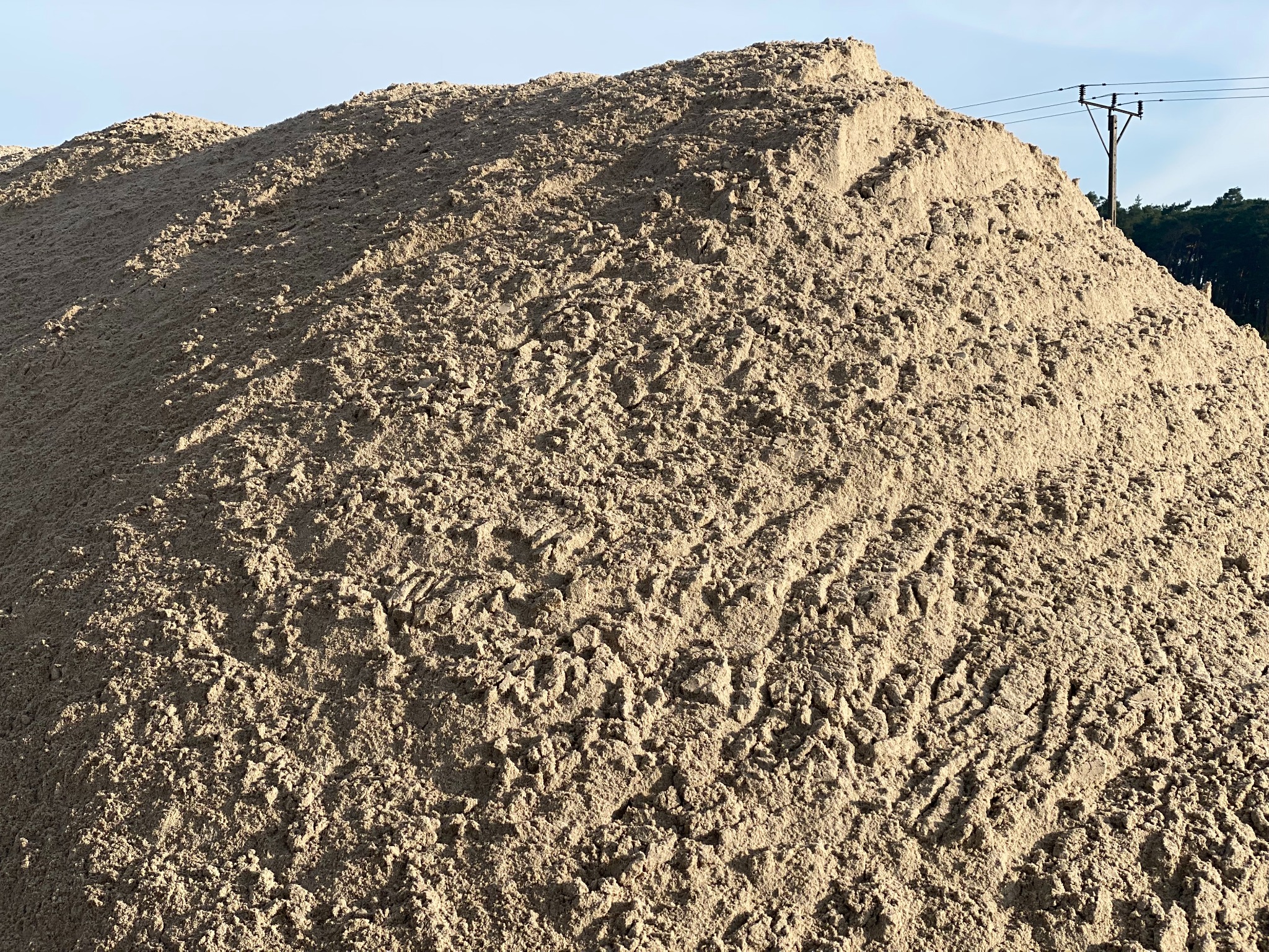 Duża pryzma piasku budowlanego o drobnej strukturze, z widocznymi śladami opon i słupem energetycznym w tle.