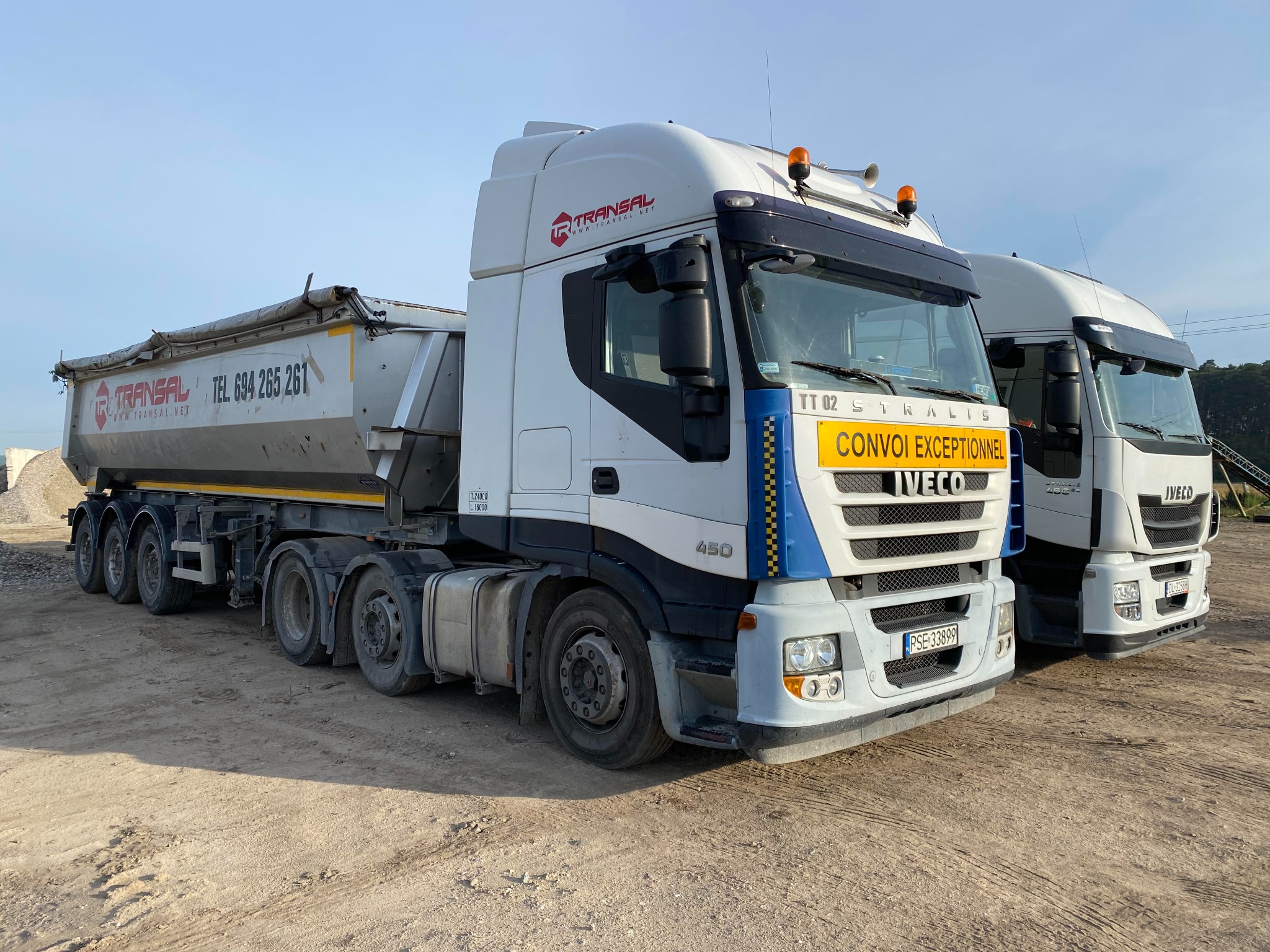 Ciężarówka Iveco Stralis 450 z naczepą, z widocznym logo firmy Transal i napisem 'Convoi Exceptionnel', zaparkowana na placu budowy obok innej ciężarówki, z niewielkim stosem kruszywa w tle.