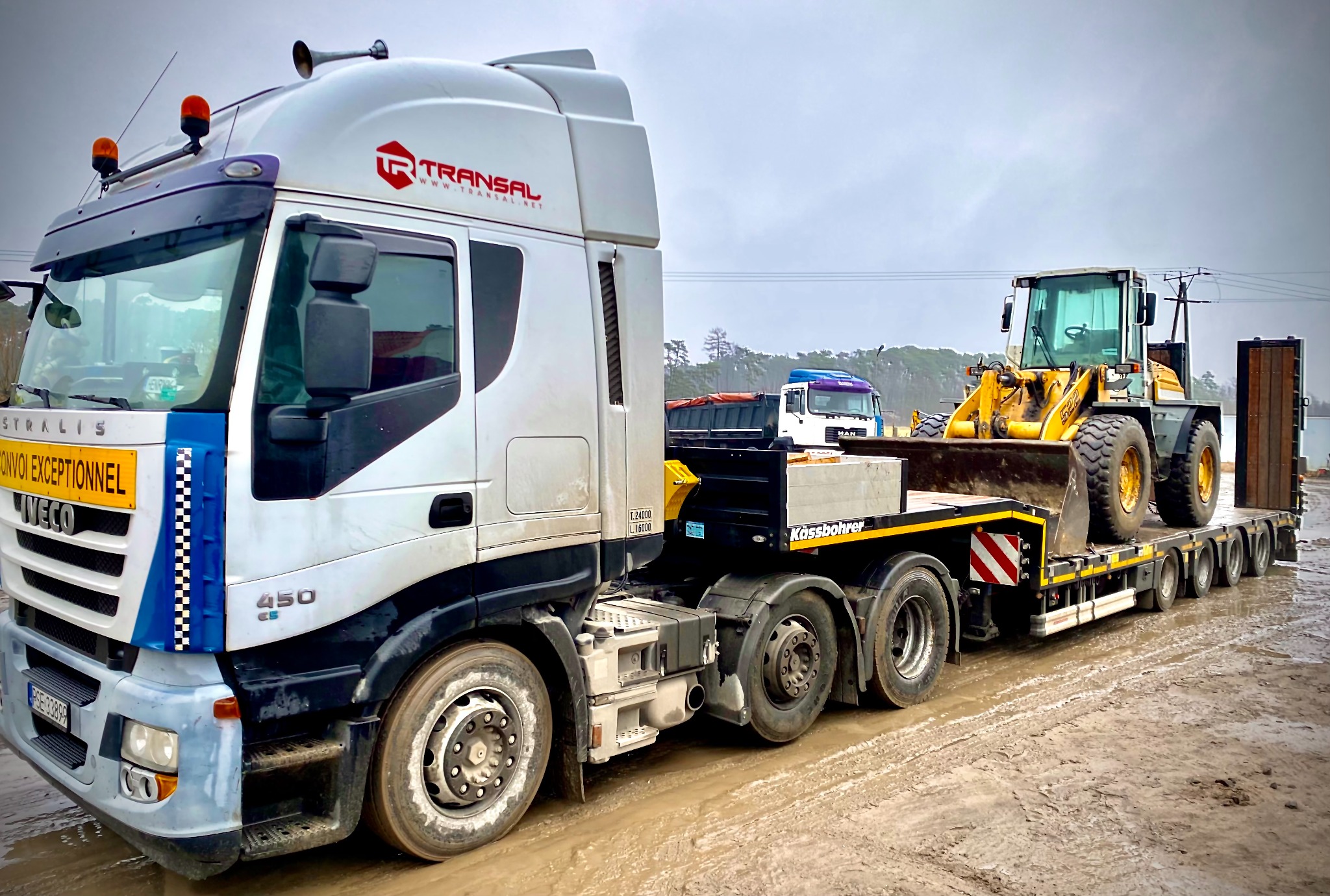 Ciężarówka laweta marki Iveco z żółtą ładowarką kołową na platformie, widoczne logo firmy transportowej Transal, w tle inne pojazdy na placu budowy.