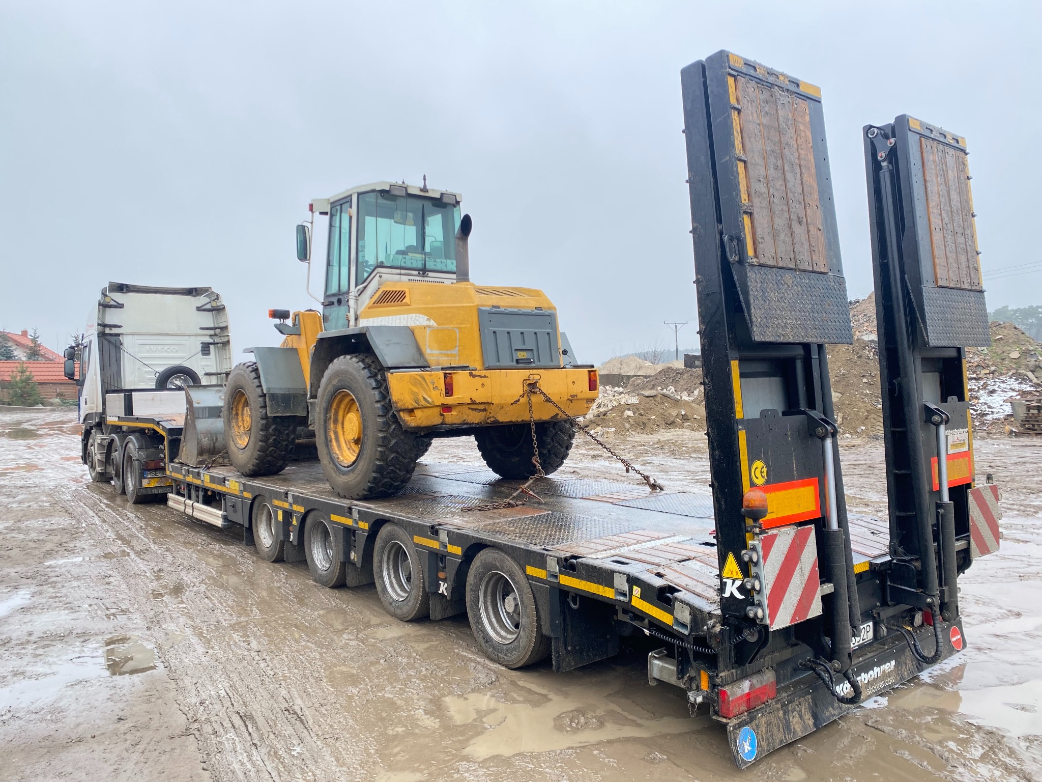 Żółta ładowarka kołowa transportowana na niskopodwoziowej przyczepie ciężarowej w pochmurny dzień, widok z tyłu na błotnistym terenie.