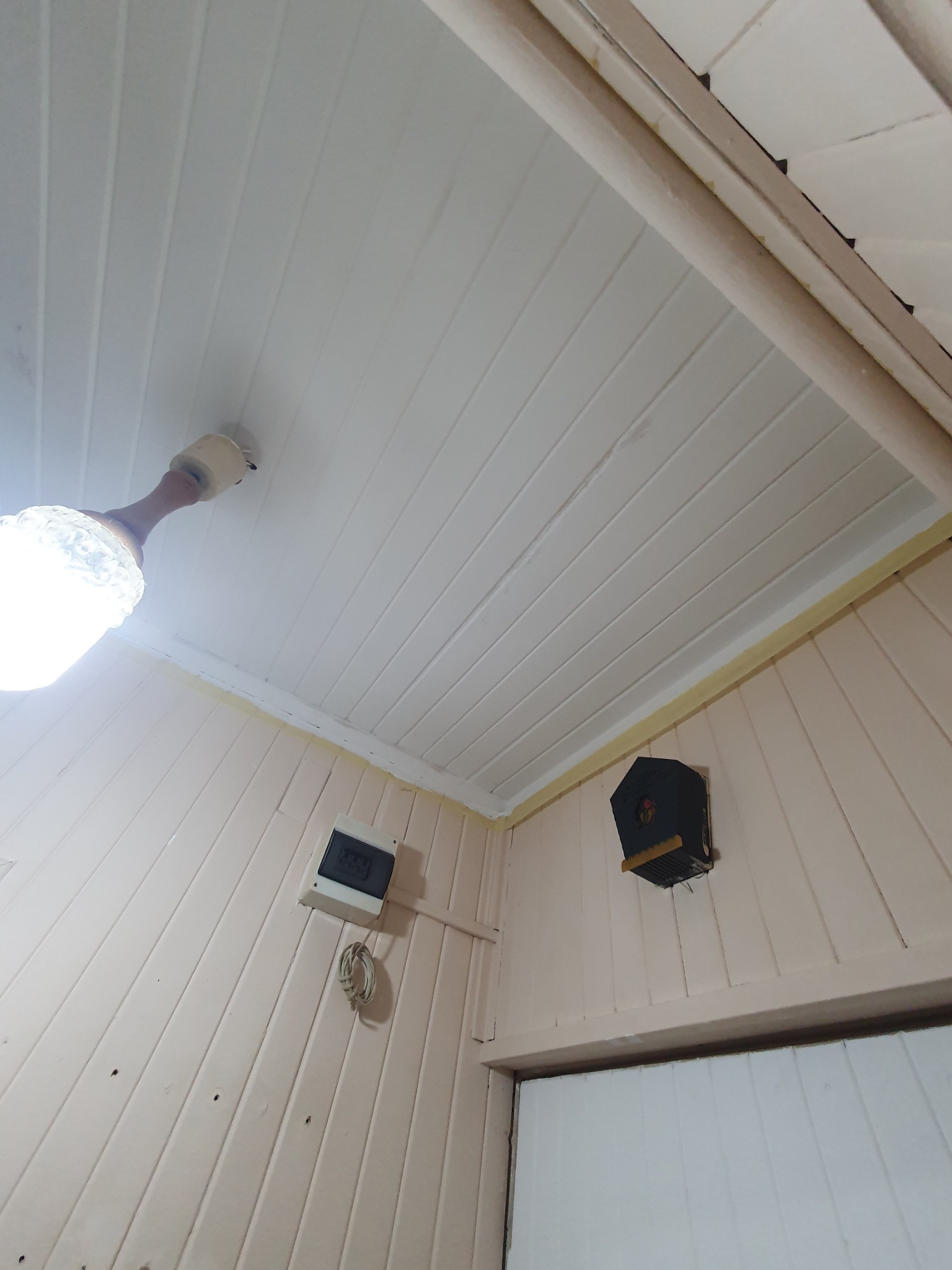 Wnętrze pomieszczenia z boazerią w kolorze pudrowego różu, biały sufit z drewnianymi panelami, wystający kabel, skrzynka elektryczna i budka dla ptaków na ścianie.