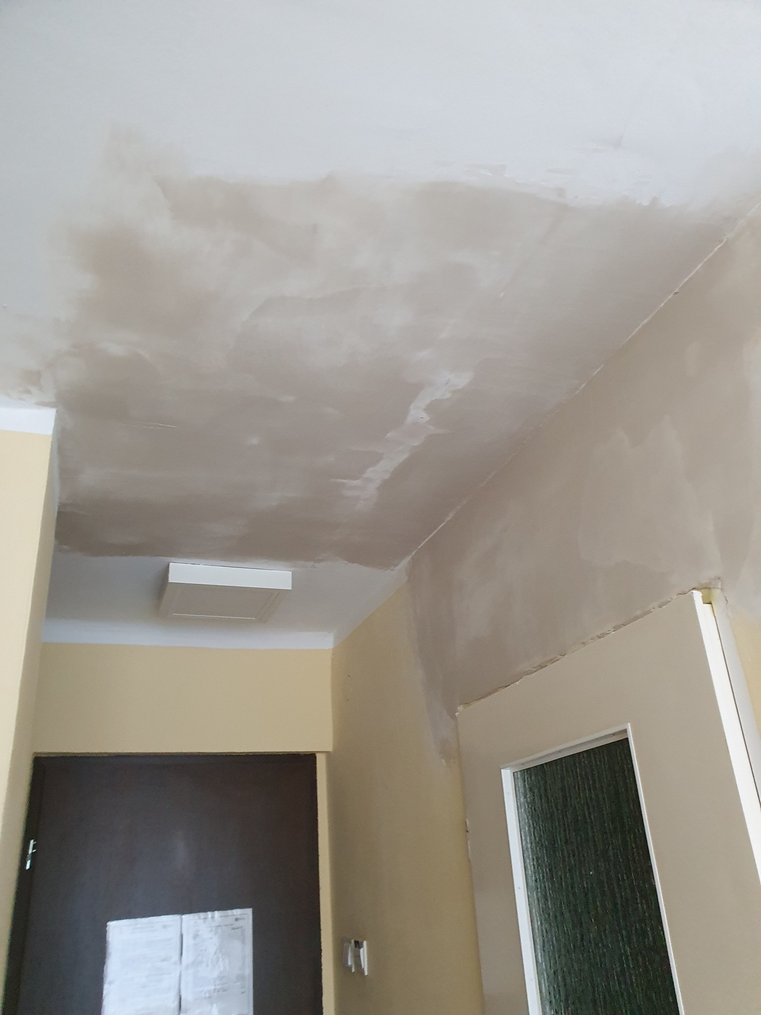 Niedokończona gładź na suficie w korytarzu, widoczne łączenia płyt gipsowych i nierówności przed szlifowaniem, ściany w odcieniach beżu, brązowe drzwi z dokumentami.