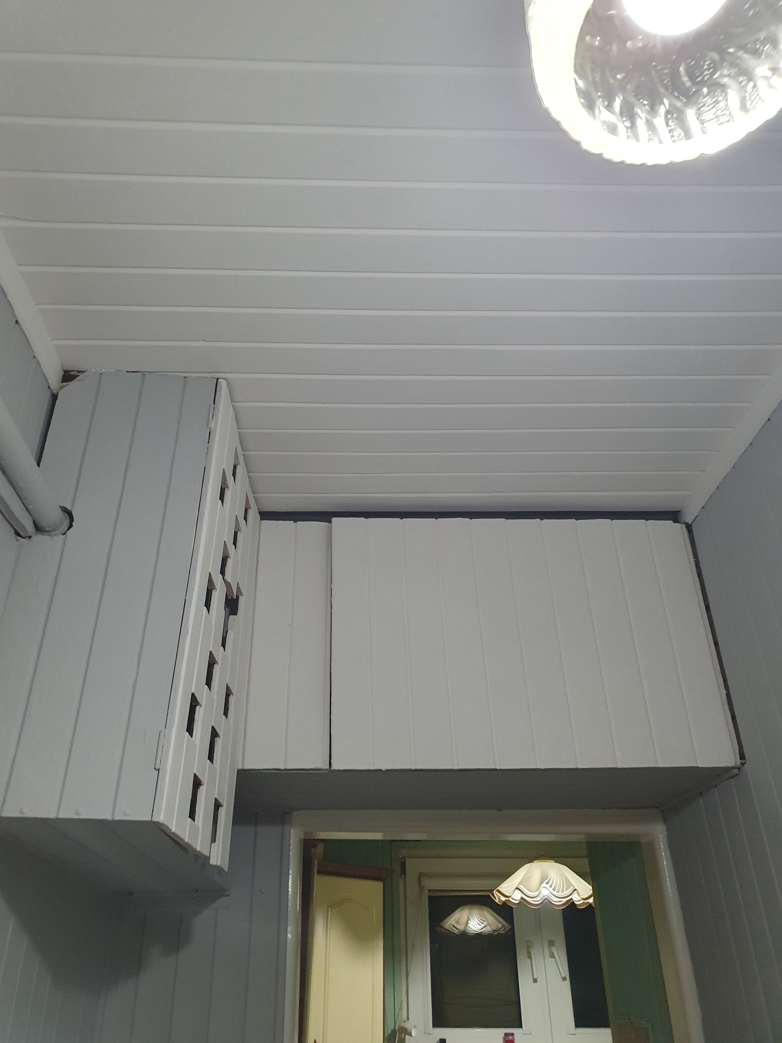 Białe panele PCV na suficie i ścianach małego pomieszczenia, widoczna lampa sufitowa i szafka z otworami wentylacyjnymi, wnętrze po remoncie.