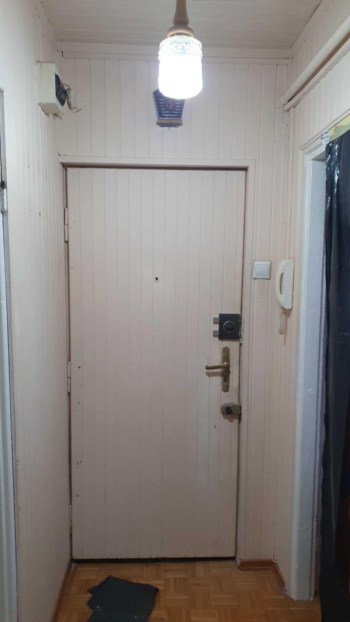 Wnętrze przedpokoju z boazerią, drewnianą podłogą i drzwiami wejściowymi z zamkiem, oświetlone lampą sufitową. Na podłodze leży czarna folia ochronna.