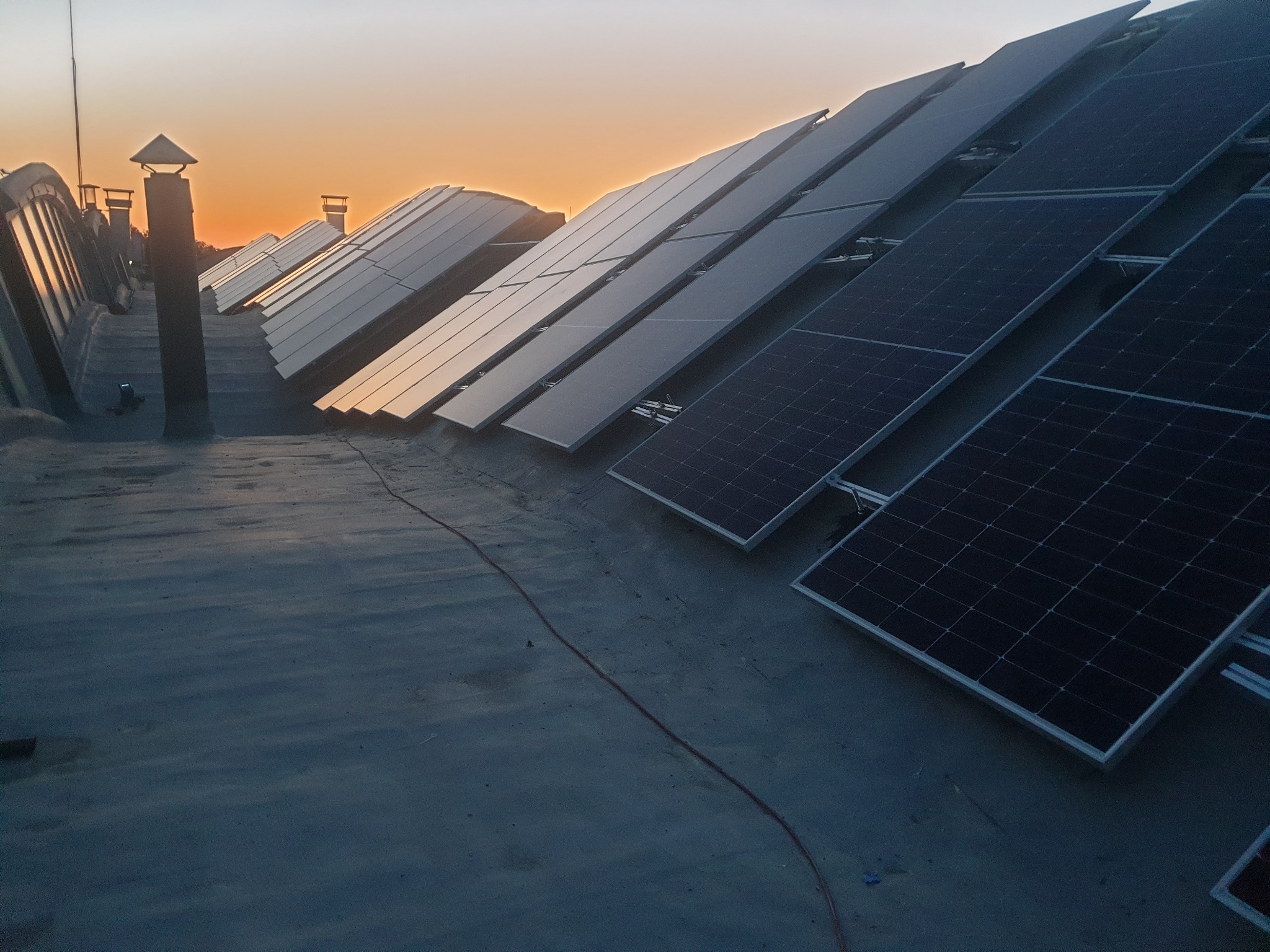 Rząd paneli słonecznych zainstalowanych na płaskim dachu w Białymstoku, sfotografowany o zmierzchu, z widocznymi kominami i okablowaniem.