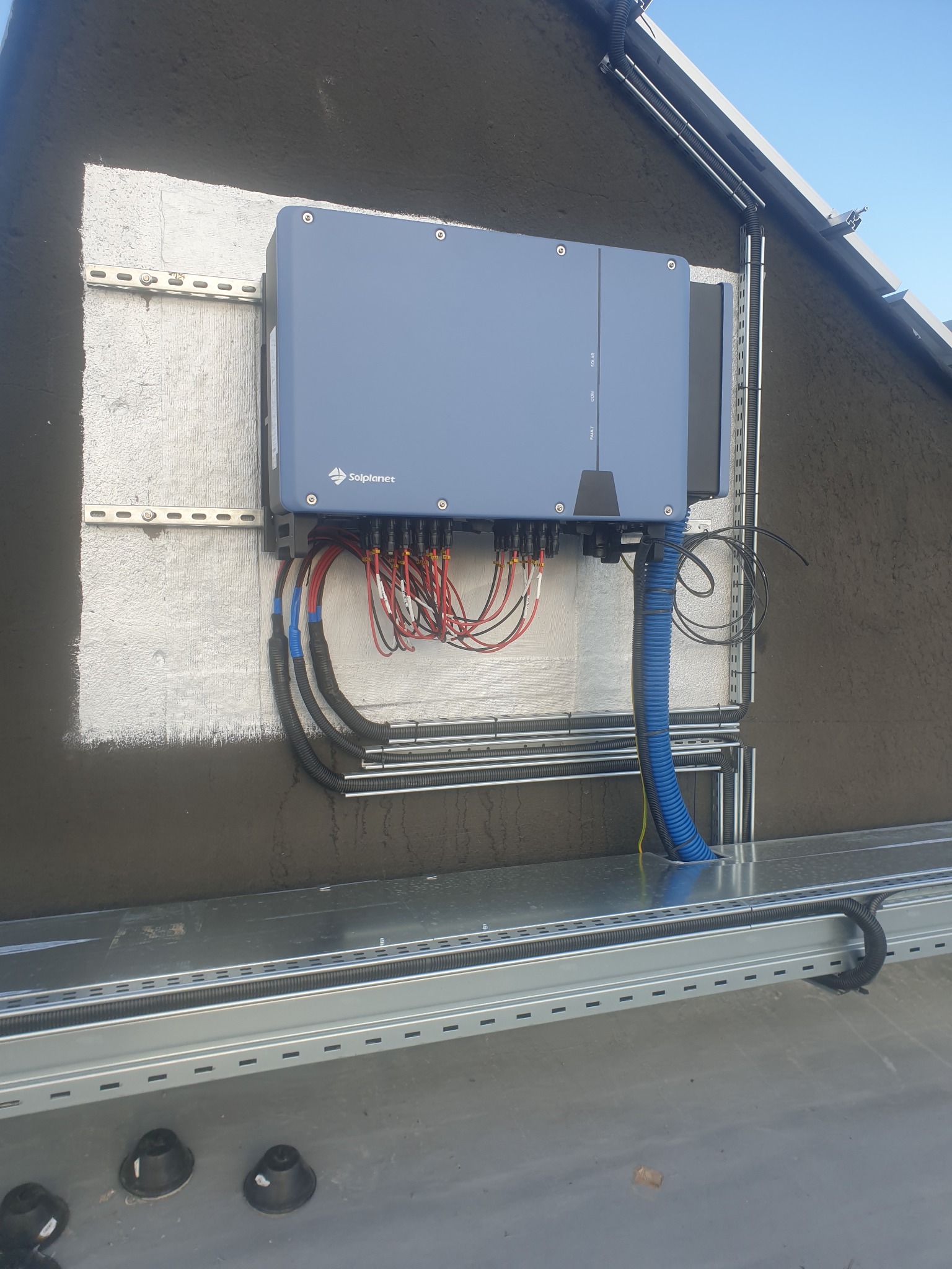 Szary inwerter Solplanet zamontowany na ścianie, z widocznymi kablami w kolorowych osłonach i metalowymi szynami montażowymi. Ściana częściowo otynkowana, z widoczną fakturą. W tle fragment dachu...