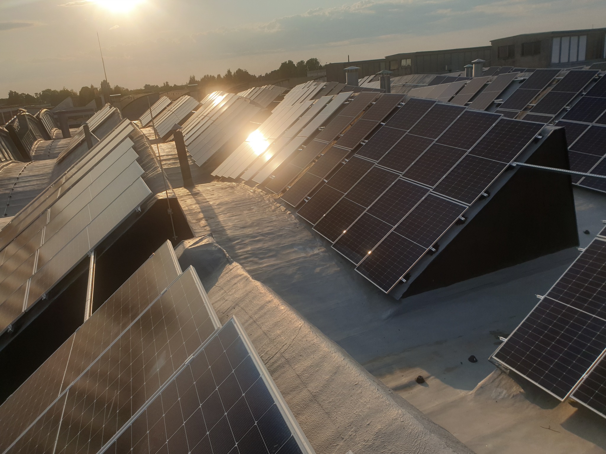 Rząd paneli słonecznych na płaskim dachu budynku przemysłowego, odbijających promienie zachodzącego słońca.