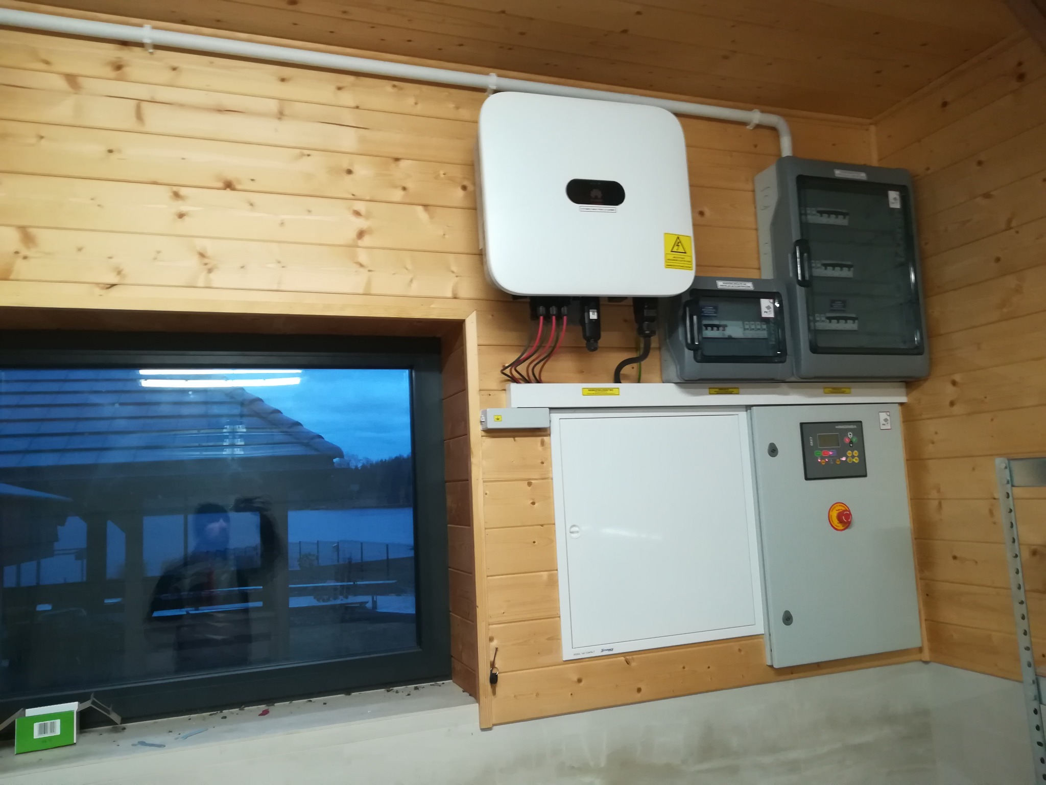Wnętrze drewnianego domu z zainstalowanym inwerterem fotowoltaicznym, rozdzielnicami elektrycznymi i widokiem na jezioro odbijającym się w oknie.