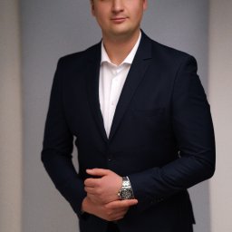 Wiesław Kozłowski Fotografia - Portret mężczyzny w ciemnym garniturze i białej koszuli, z rękami splecionymi i widocznym zegarkiem na nadgarstku na neutralnym tle.