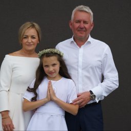 Wiesław Kozłowski Fotografia - Dziewczynka w białej sukience i wianku na głowie, stojąca z rodzicami na tle ciemnej ściany, ręce złożone do modlitwy, widoczny srebrny medalik.