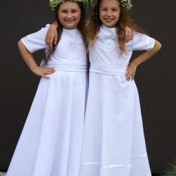 Wiesław Kozłowski Fotografia - Dwie uśmiechnięte dziewczynki w białych albach i wiankach na głowach, pozujące na tle ciemnej ściany.