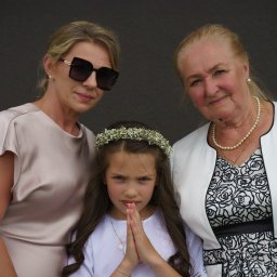 Wiesław Kozłowski Fotografia - Trzy pokolenia kobiet: dziewczynka w wianku z gipsówki, jej mama w sukience w odcieniu szampana i babcia w białym żakiecie z perłami, pozują razem na tle ciemnej ściany.