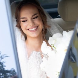 Wiesław Kozłowski Fotografia - Uśmiechnięta panna młoda w sukni z koronkowymi zdobieniami i welonem, siedząca w samochodzie z bukietem białych orchidei.