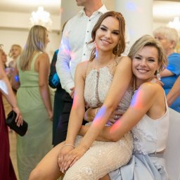 Wiesław Kozłowski Fotografia - Dwie uśmiechnięte kobiety w sukienkach wieczorowych, siedzące obok siebie na tle gości weselnych i dekoracji świetlnych.