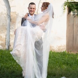 Wiesław Kozłowski Fotografia - Uśmiechnięty pan młody trzyma na rękach pannę młodą w białej sukni i welonie na tle starej ściany z roślinnością.