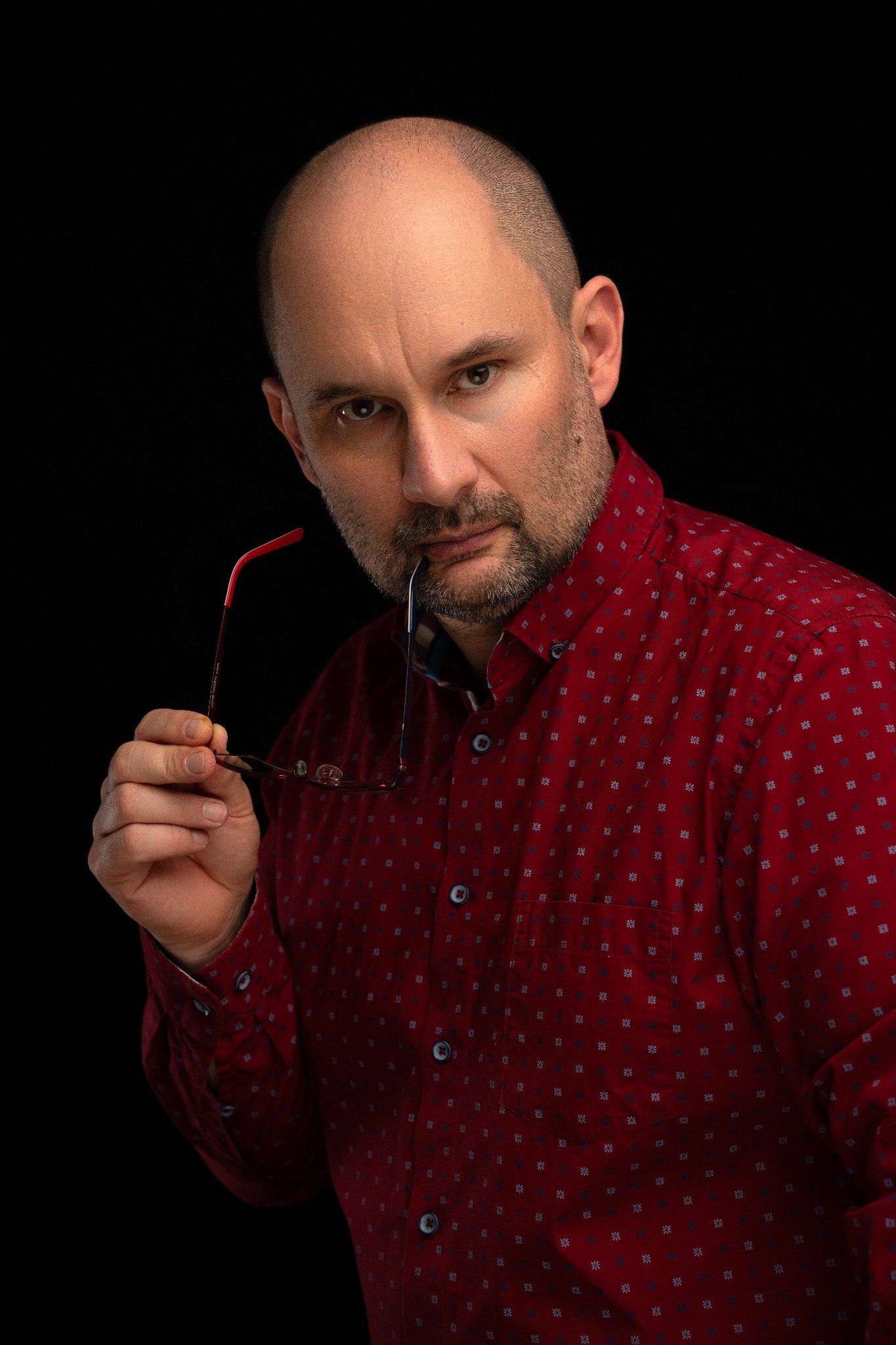 Portret mężczyzny w czerwonej koszuli z drobnym wzorem, trzymającego okulary z czerwonymi zausznikami w ustach na czarnym tle.