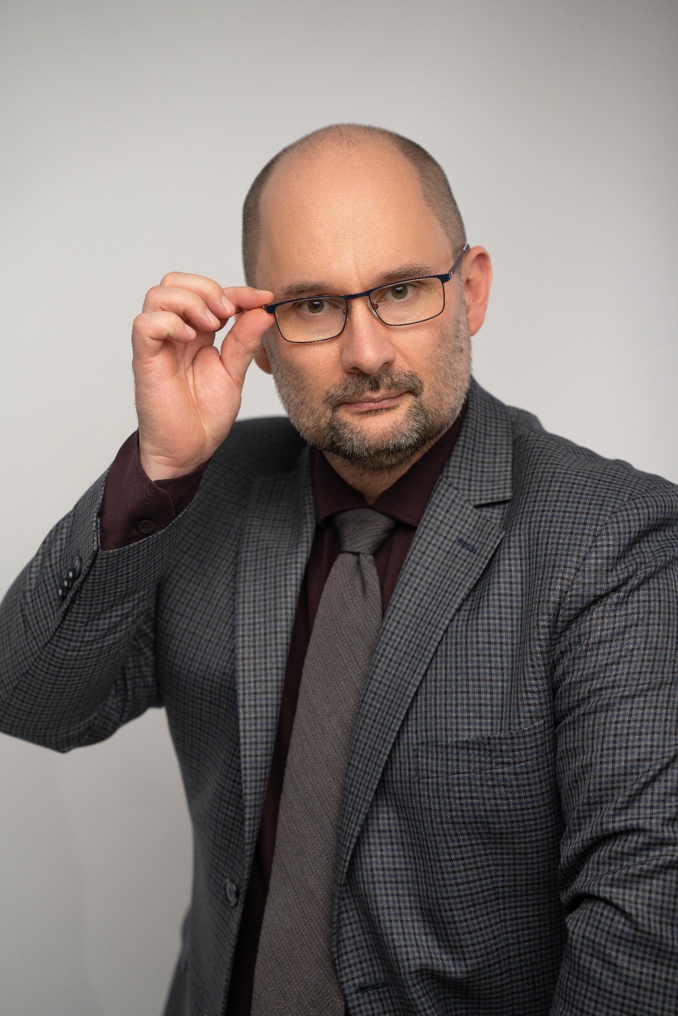 Portret mężczyzny w szarej marynarce w kratę, bordowej koszuli i szarym krawacie, poprawiającego okulary na tle jasnoszarego tła.
