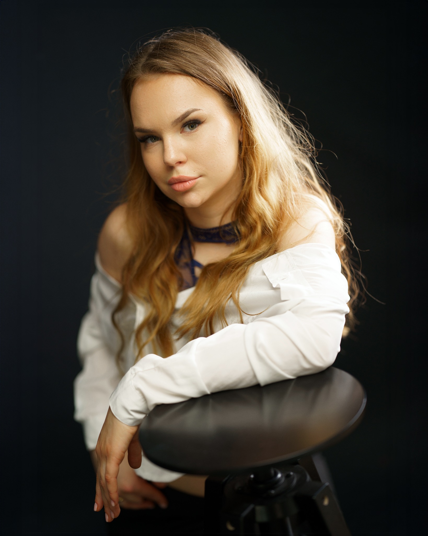 Portret młodej kobiety z długimi blond włosami, ubrana w białą koszulę z odkrytymi ramionami i koronkowy choker, opierająca się o czarny taboret na ciemnym tle.