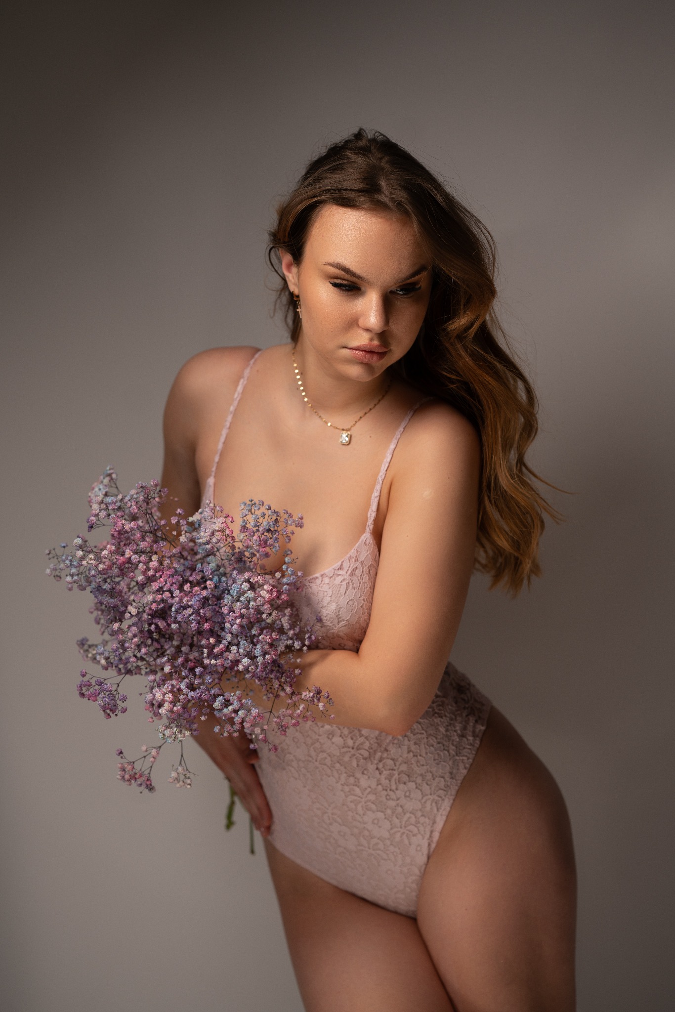 Młoda kobieta w pudrowym body z koronki pozuje z bukietem gipsówki w odcieniach różu i fioletu, na szyi złoty łańcuszek, włosy opadają na ramię.