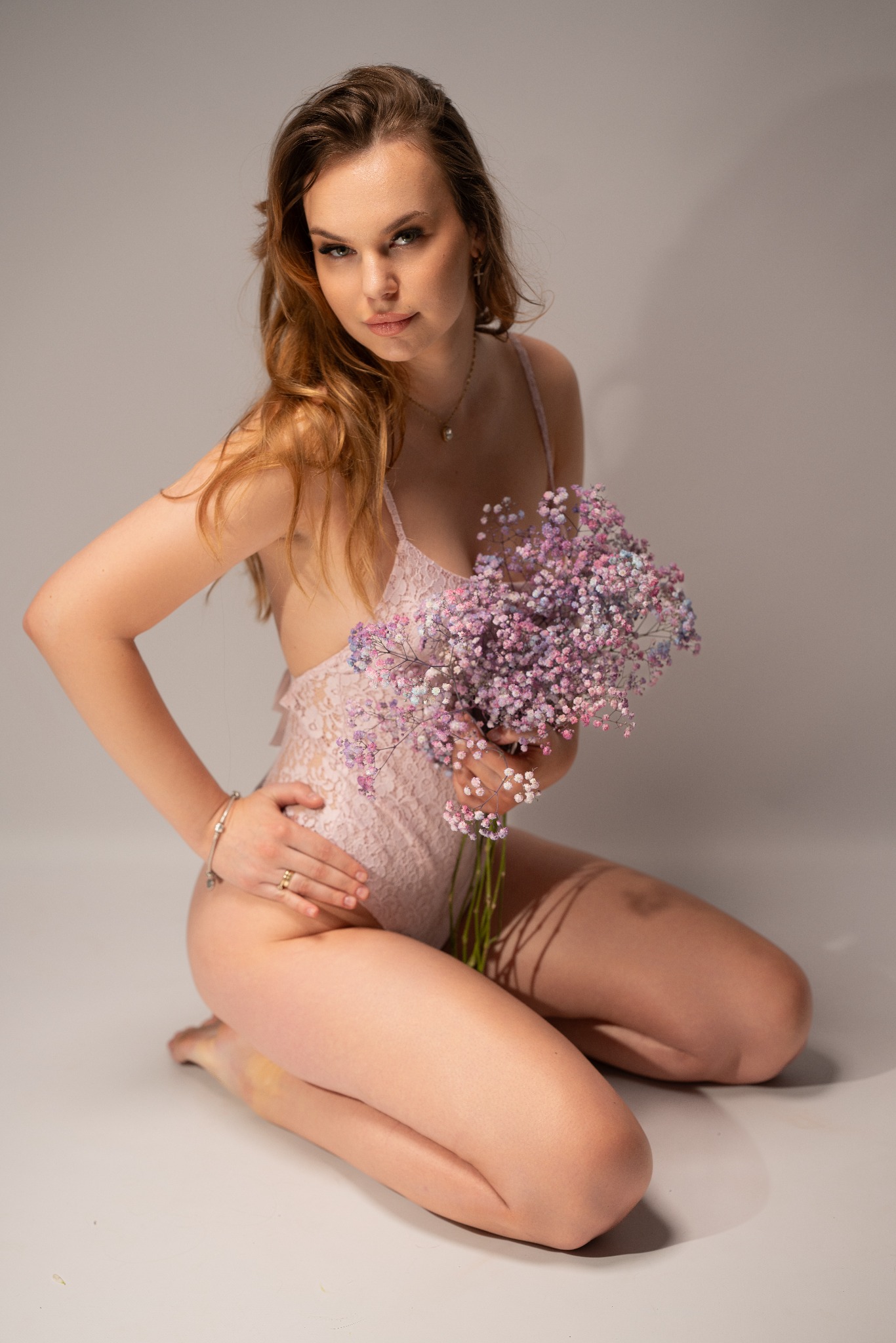 Kobieta w koronkowym body, klęcząca na szarym tle, trzyma bukiet pastelowych kwiatów gipsówki, pozuje z delikatnym uśmiechem.