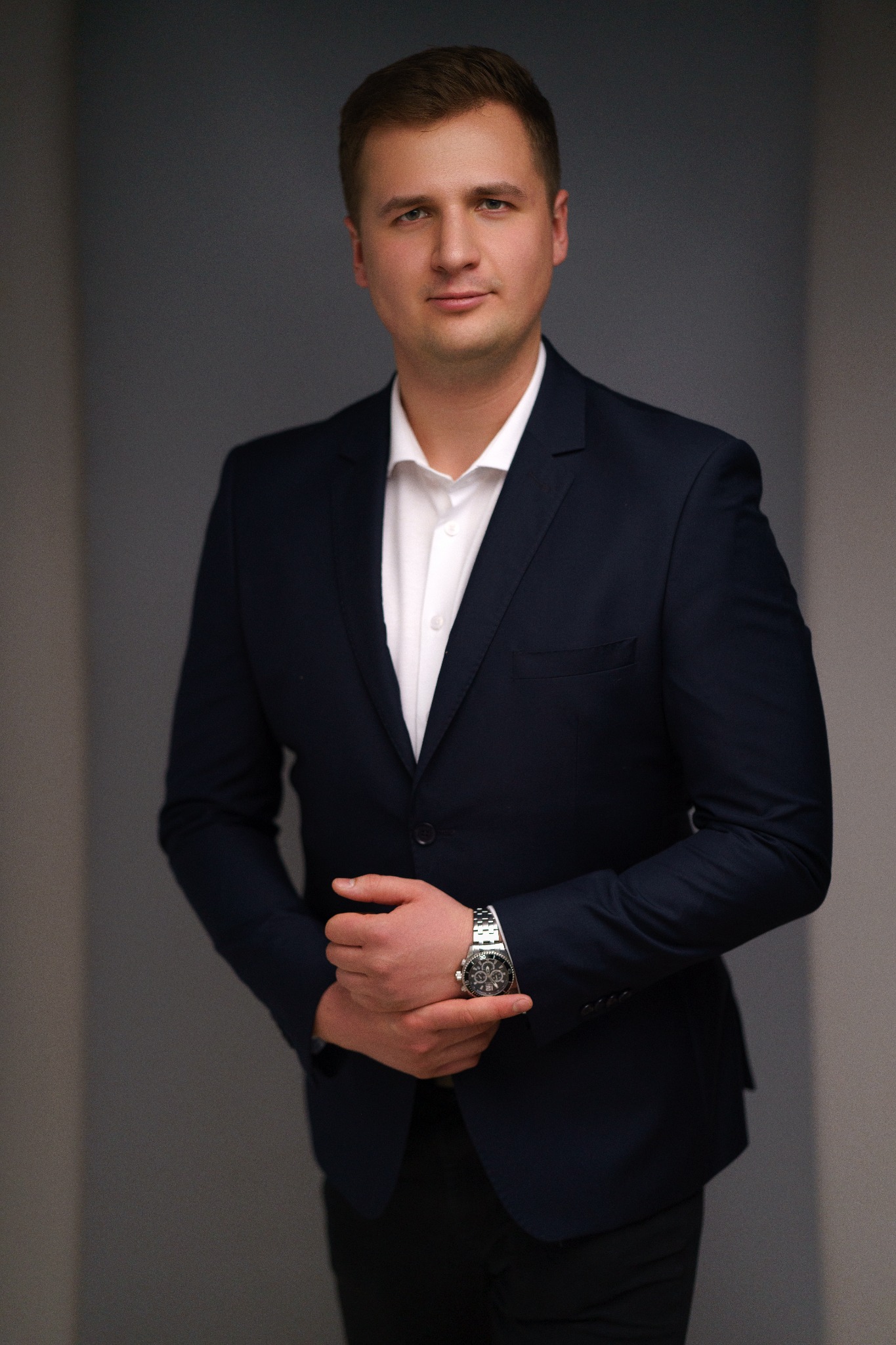 Mężczyzna w granatowym garniturze i białej koszuli, z zegarkiem na ręce, pozuje na neutralnym tle.