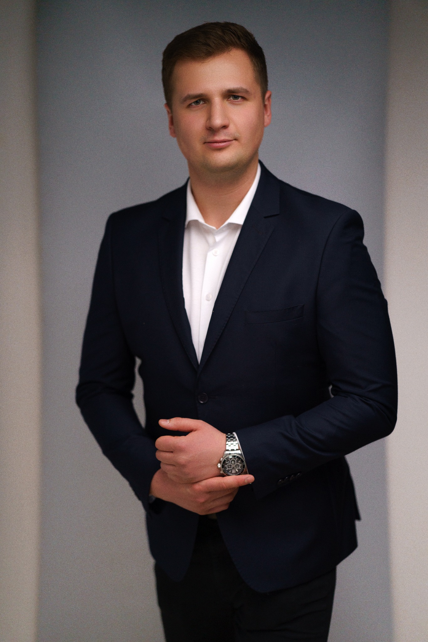 Portret mężczyzny w ciemnym garniturze i białej koszuli, z rękami splecionymi i widocznym zegarkiem na nadgarstku na neutralnym tle.