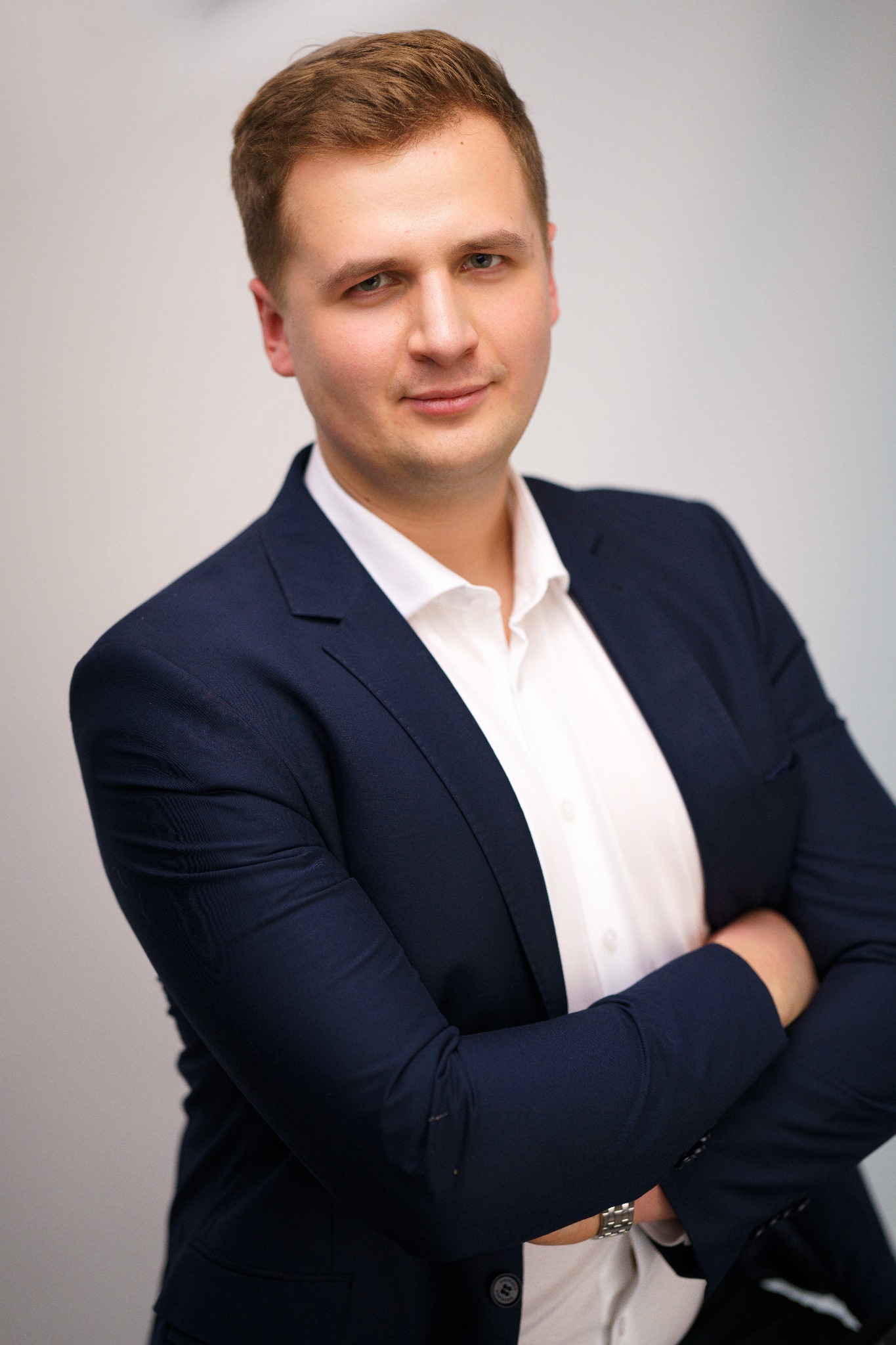 Portret młodego mężczyzny w granatowej marynarce i białej koszuli, z założonymi rękami na neutralnym tle.