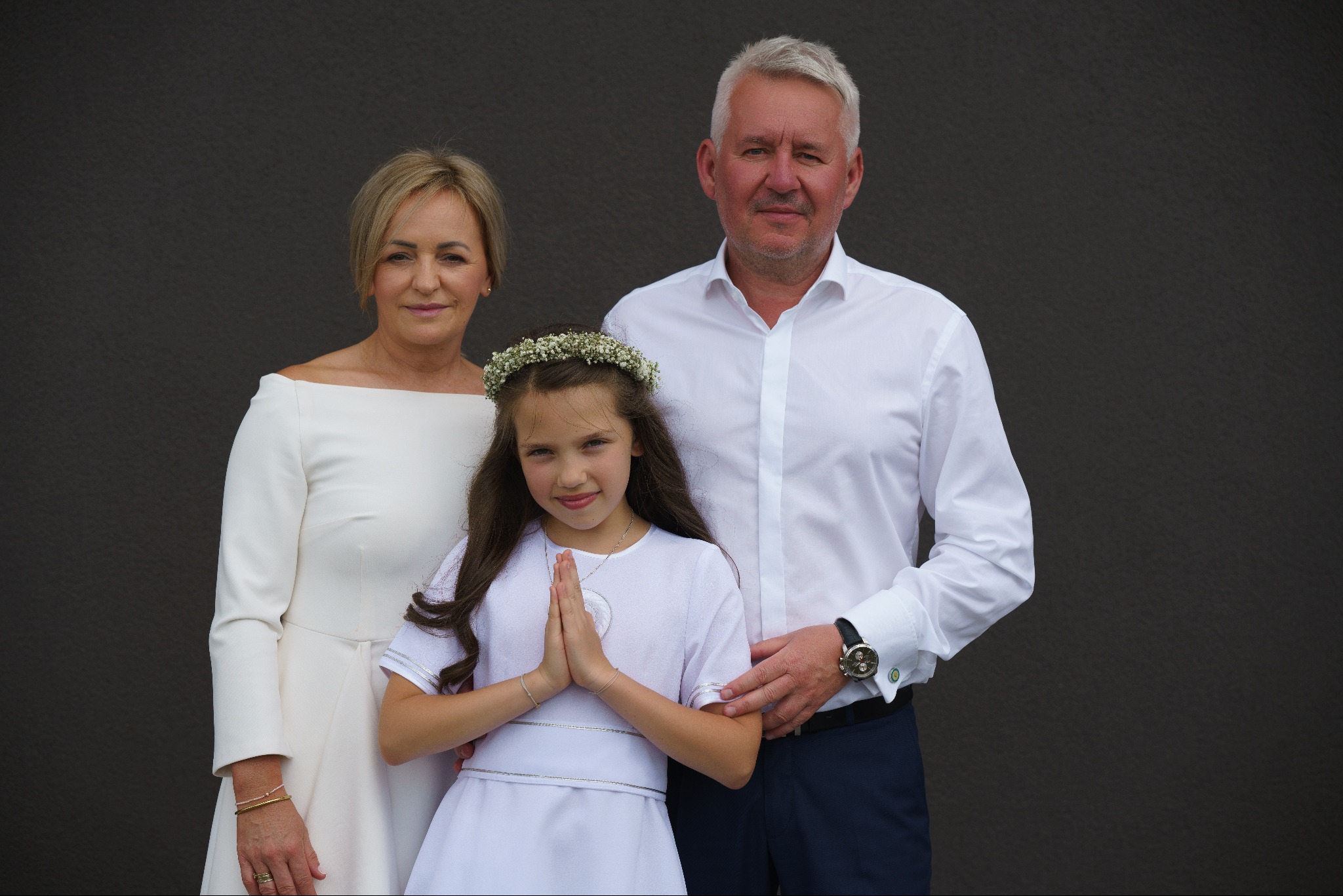 Dziewczynka w białej sukience i wianku na głowie, stojąca z rodzicami na tle ciemnej ściany, ręce złożone do modlitwy, widoczny srebrny medalik.