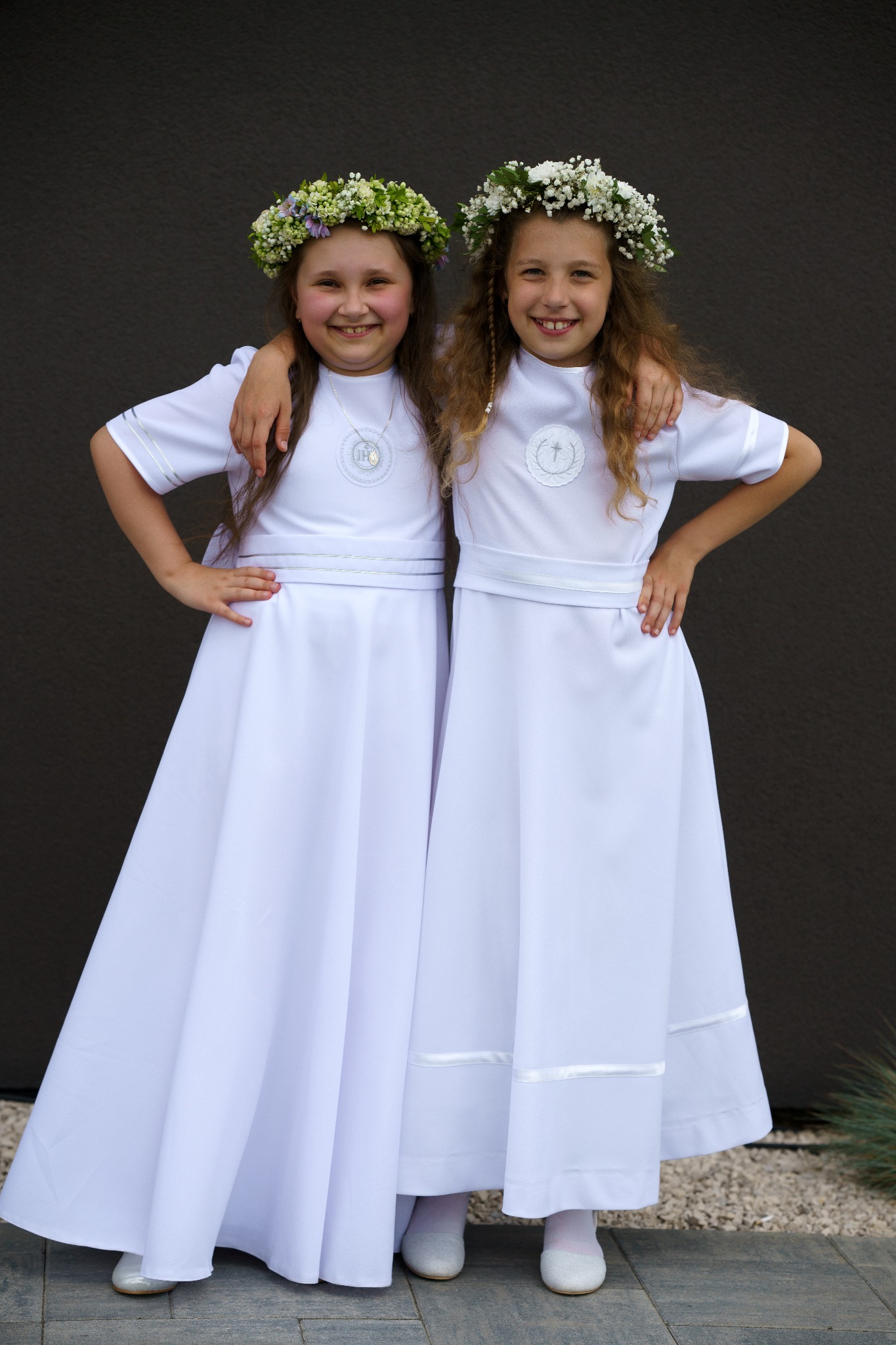 Dwie uśmiechnięte dziewczynki w białych albach i wiankach na głowach, pozujące na tle ciemnej ściany.