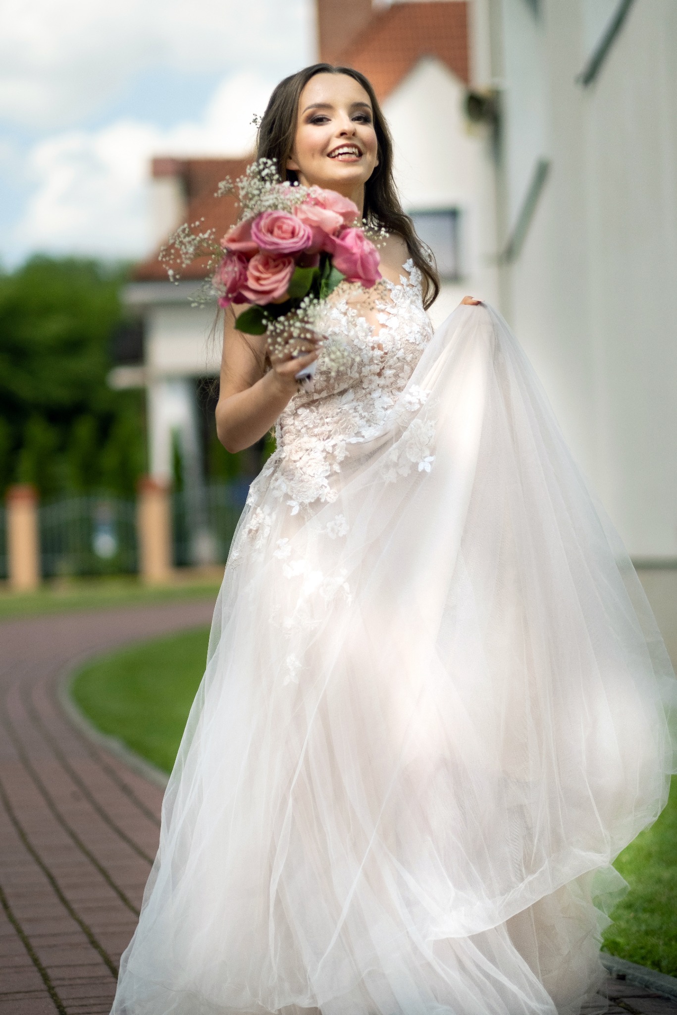 Uśmiechnięta panna młoda w koronkowej sukni z tiulowym dołem, trzymająca bukiet różowych róż i gipsówki, pozująca na tle budynku w jasnym kolorze.